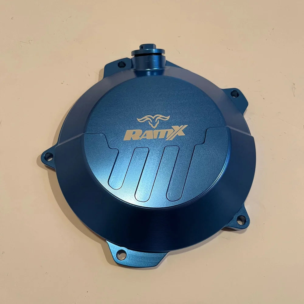 A blue anodized metal motorcycle engine cover with a gold RAMX logo, photographed from a top-down angle on a beige background.